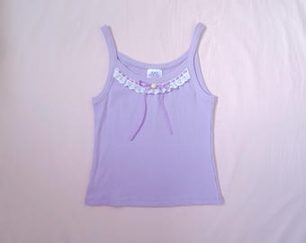 May include: A lavender ribbed tank top with spaghetti straps. The top features a decorative neckline with white lace trim, small yellow and purple accents, and two lavender ribbons. The tank top is a light purple color.