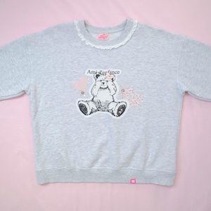 Pode incluir: Uma camisola cinzenta clara de manga comprida com um rebordo de renda branca à volta do decote redondo. O gráfico central apresenta uma ilustração a preto e branco de um urso de peluche sentado a tapar os olhos, com o texto Ami d'enfance.