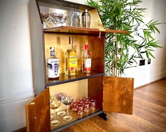 May include: A wooden bar cabinet with a hinged top and doors, featuring shelves stocked with glassware, bottles of liquor, and a cocktail shaker. The cabinet has a dark exterior with a gold interior.