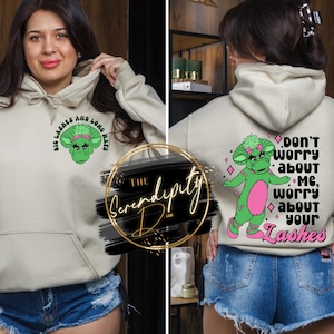 Puede incluir: Sudadera con capucha beige con bolsillo delantero y capucha con cordón. La parte delantera presenta un personaje de dibujos animados verde con el texto "BIG LASHES AND LONG NAILS". La parte trasera tiene un personaje a juego y el texto "DON'T WORRY ABOUT ME, WORRY ABOUT YOUR LASHES".
