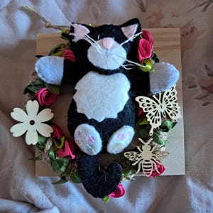 Pode incluir: Um gato de feltro preto e branco com orelhas e nariz cor-de-rosa está sentado em uma coroa feita de rosas cor-de-rosa, folhas verdes e barbante marrom. A coroa está sobre um fundo de madeira. Há uma borboleta de madeira e uma abelha de madeira na coroa.