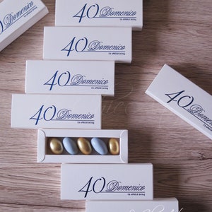 Può includere: Scatole rettangolari bianche con testo blu "40 Domenico 13 APRILE 2024". Una scatola aperta mostra caramelle dorate e azzurre. Le scatole sono disposte su una superficie di legno chiaro.