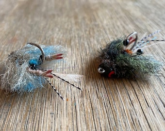 May include: Two fly fishing lures, one with a blue and white body and the other with a green and brown body. Both lures have black and white feathers.