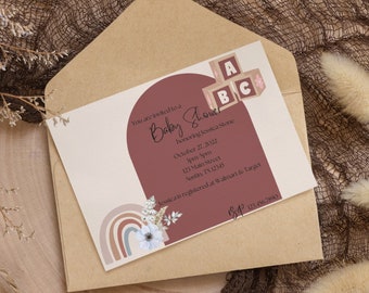 May include: A baby shower invitation with a brown envelope and a cream card with a pink and brown circle with the text "You are invited to a Baby Shower honoring Jessica Stone" and the date and time of the event. The card also has a rainbow with white flowers and a pink, brown, and white block with the letters "ABC" on it.