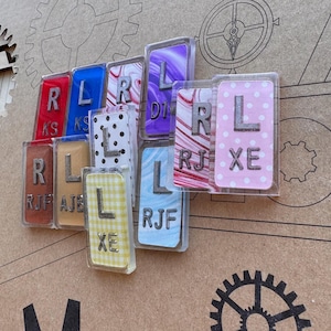 May include: A collection of rectangular, clear acrylic magnets with various colorful backgrounds and silver letters. The magnets display the letters "R", "L", and "XE", with some featuring additional text. The backgrounds include polka dots, stripes, and plaid patterns.