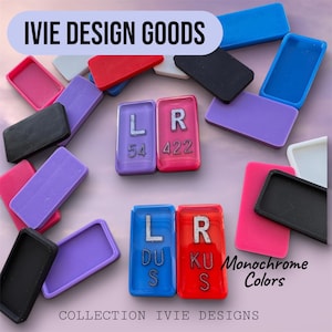 May include: A collection of rectangular plastic tiles in various colors including red, blue, purple, black, pink, and white. Some tiles feature silver letters and numbers such as L 54, R 422, L DUS, and R KUS. Text reads IVIE DESIGN GOODS and Monochrome Colors.
