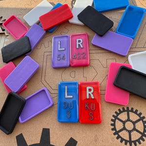 May include: A collection of colorful rectangular game pieces, including black, red, blue, purple, pink, and white. Two sets of game pieces have the letters "LR" with numbers and letters. The pieces are scattered on a brown surface.