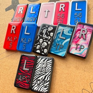 May include: A collection of rectangular resin pieces with various designs and letters. Some have glittery letters, others feature patterns like zebra stripes, skulls, and marbled effects. Colors include red, blue, pink, and black. The letters include R, L, and other text.