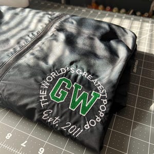 May include: Black and green embroidered jacket with the text "The World's Greatest Pop Pop" and "Est. 2011" in a circle.