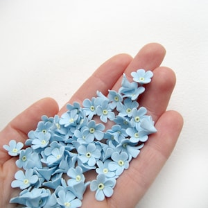 May include: A hand holding a collection of light blue forget-me-not flower embellishments.