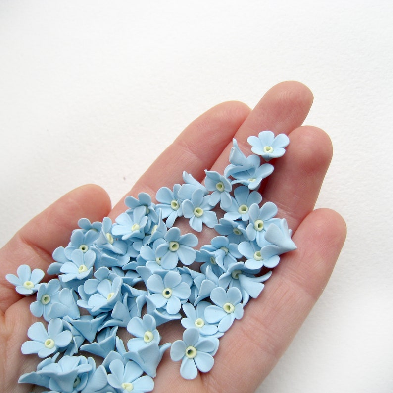 May include: A hand holding a collection of light blue forget-me-not flower embellishments.