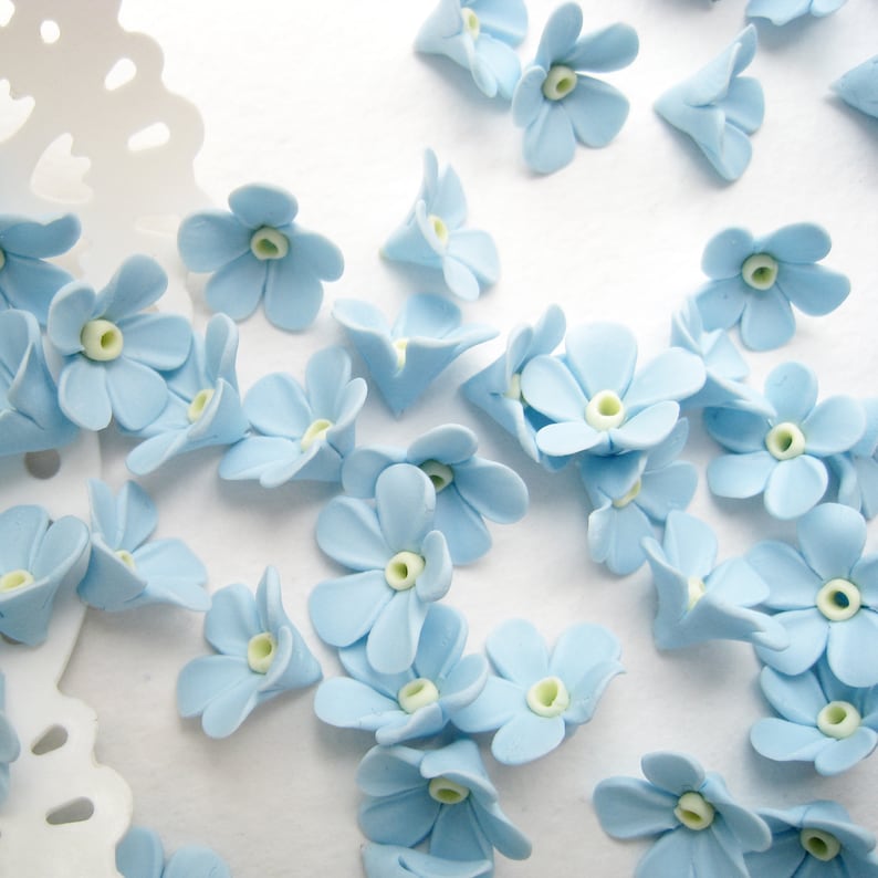 May include: A collection of light blue forget-me-not flower embellishments. The flowers are made of a soft, pliable material and have a delicate, realistic appearance.