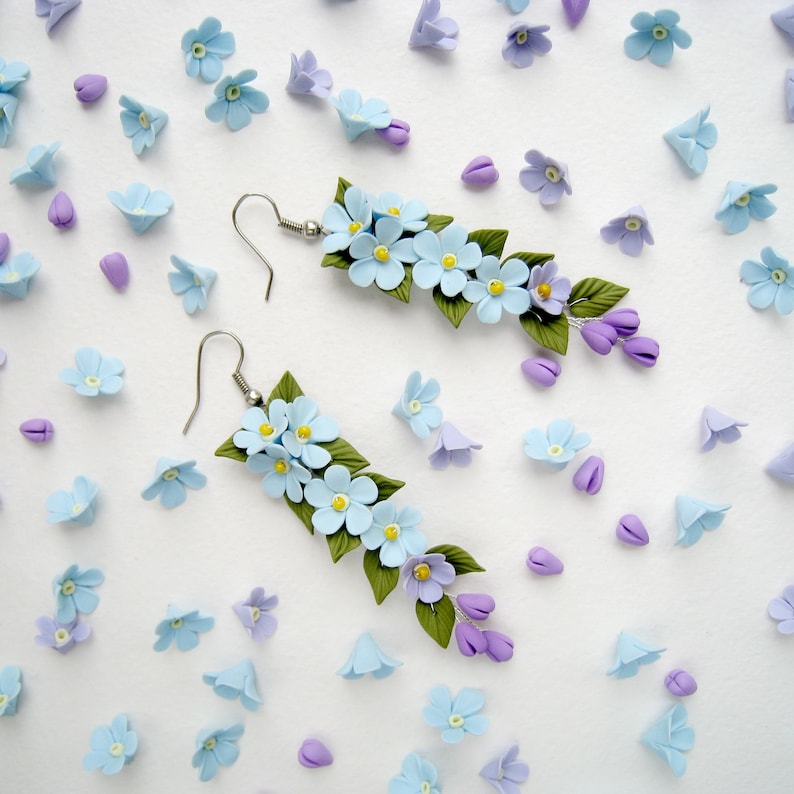 May include: A pair of dangle earrings featuring blue and purple polymer clay flowers. The earrings are made with silver-toned hooks.