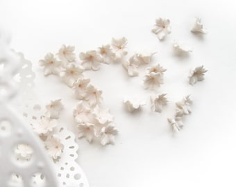May include: A collection of delicate, off-white porcelain flowers scattered on a white background. The flowers are arranged in a loose, organic pattern, creating a sense of whimsy and elegance.