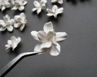 May include: A collection of white porcelain flowers, some of which are being held by a pair of tweezers. The flowers are arranged on a grey surface.
