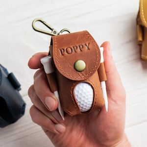 May include: Three leather golf ball holders in navy blue, brown, and tan. Each holder has a snap closure and a metal clip. The brown holder has the name "POPPY" embossed on it, and the tan holder has "AVA". The brown holder also holds a golf ball and tees.