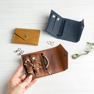 May include: Three small leather jewelry organizers in tan, blue, and brown. The tan case is closed with the name EVY embossed on the front. The blue and brown cases are open, revealing interior slots for earrings, rings, and a zippered pocket.