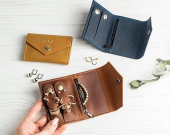 Può includere: Tre piccoli organizzatori di gioielli in pelle color cuoio, blu e marrone. La custodia color cuoio è chiusa con il nome EVY impresso sul davanti. Le custodie blu e marrone sono aperte, rivelando scomparti per orecchini, anelli e una tasca con cerniera.
