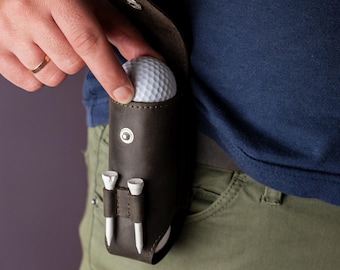 May include: A dark brown leather golf ball holder with two white golf tees. The holder is attached to a person's waist. A white golf ball is partially visible. The person is wearing a blue shirt and green pants.