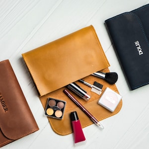 Puede incluir: Vista cenital de tres estuches de cosméticos de cuero en marrón, beige y azul marino. El estuche beige está abierto, mostrando brochas de maquillaje, pintalabios y una paleta de sombras de ojos. El estuche azul marino tiene el nombre "DOLIE" en relieve, y el marrón "CAMBER".