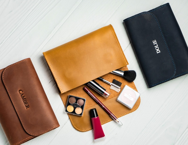 May include: An overhead shot of three leather cosmetic cases in brown, tan, and navy blue. The tan case is open, revealing makeup brushes, lipsticks, and an eyeshadow palette. The navy case has the name "DOLIE" embossed on it, and the brown case has "CAMBER" embossed.