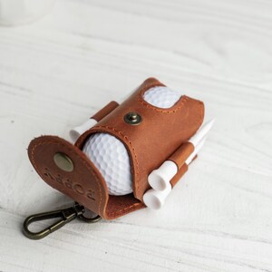 May include: A brown leather golf ball holder with a snap closure, a metal clasp, and the word "POPPY" embossed on the flap. The holder contains a white golf ball and several white golf tees.