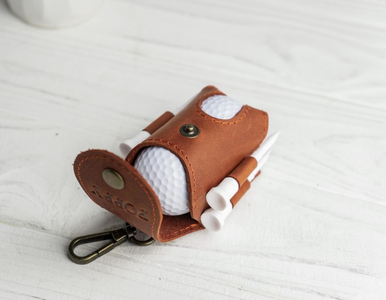 May include: A brown leather golf ball holder with a snap closure, a metal clasp, and the word "POPPY" embossed on the flap. The holder contains a white golf ball and several white golf tees.