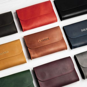 May include: An assortment of leather wallets in various colors, including black, red, brown, and blue. Some wallets have embossed names like "DOLIE" and "CAMBER". The wallets have a simple, rectangular design with a flap closure.
