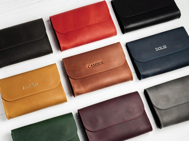 May include: An assortment of leather wallets in various colors, including black, red, brown, and blue. Some wallets have embossed names like "DOLIE" and "CAMBER". The wallets have a simple, rectangular design with a flap closure.