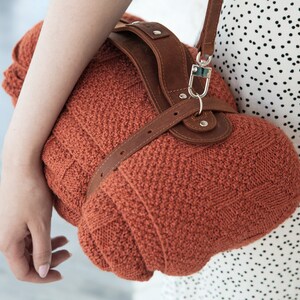 May include: A rust-colored, knitted handbag with a brown leather strap and accents. The bag has a rounded shape and a textured knit pattern. The strap is attached with silver-colored hardware.