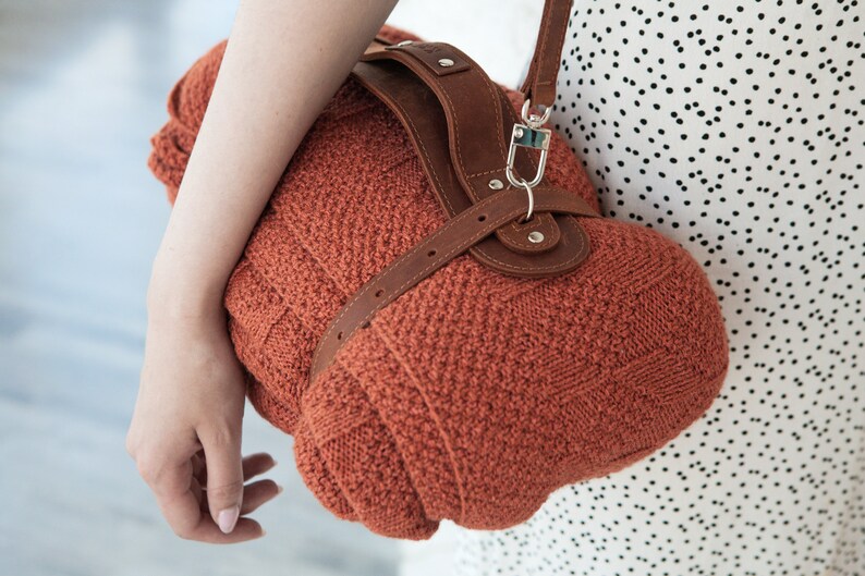 May include: A rust-colored, knitted handbag with a brown leather strap and accents. The bag has a rounded shape and a textured knit pattern. The strap is attached with silver-colored hardware.