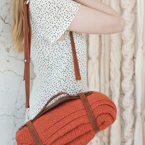 May include: An orange knitted blanket rolled and secured with brown leather straps, carried over the shoulder with a matching leather strap. The blanket has a textured appearance. The bag is attached with silver metal clasps.