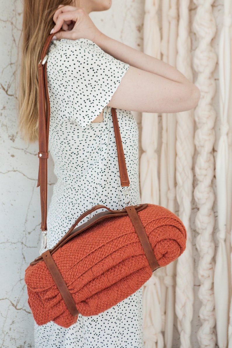May include: An orange knitted blanket rolled and secured with brown leather straps, carried over the shoulder with a matching leather strap. The blanket has a textured appearance. The bag is attached with silver metal clasps.