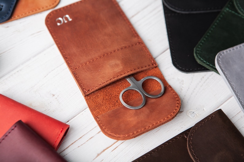 May include: A brown leather case with a small pair of scissors inside. The case has a flap that closes with a snap. The case is monogrammed with the initials "UC".