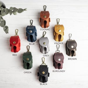 May include: A collection of leather AirPod cases in various colors, including red, blue, green, and brown. Each case has a snap closure, a metal clasp, and a name or word embossed on it. The cases are displayed on a light wood surface.