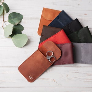 May include: A set of eight leather pouches in various colors, including brown, red, burgundy, gray, black, and navy blue. The pouches have a flap closure and are arranged in a semi-circle on a white wooden surface. One pouch has a pair of silver scissors inside and the initials "UC" embossed on the front.