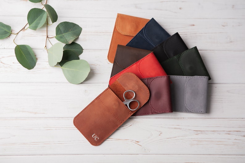 May include: A set of eight leather pouches in various colors, including brown, red, burgundy, gray, black, and navy blue. The pouches have a flap closure and are arranged in a semi-circle on a white wooden surface. One pouch has a pair of silver scissors inside and the initials "UC" embossed on the front.