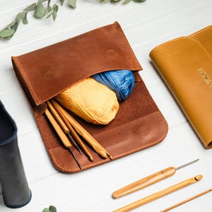 May include: A brown leather roll-up case containing yellow and blue yarn balls and wooden crochet hooks. A second closed mustard yellow leather case with the name ALYSON embossed on the front and a dark blue leather case are also visible.
