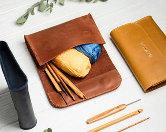 May include: A brown leather case with a flap closure, containing yarn and knitting needles. The case is open, revealing the yarn and needles inside. There is also a smaller, yellow leather case with the word "ALYX" embossed on it.