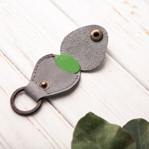 May include: A gray leather guitar pick holder with a green pick inside. The holder has a metal ring for attaching to a keychain.