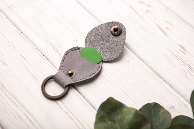 May include: A gray leather guitar pick holder with a green pick inside. The holder has a metal ring for attaching to a keychain.