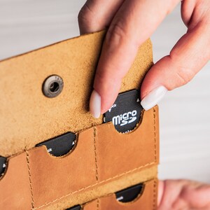 K&ouml;nnte beinhalten: Ein braunes Lederetui mit mehreren Steckpl&auml;tzen zum Aufbewahren von Speicherkarten. Eine Hand h&auml;lt eine Micro-SD-Karte mit der Aufschrift "micro SD" und steckt sie in einen der Steckpl&auml;tze.
