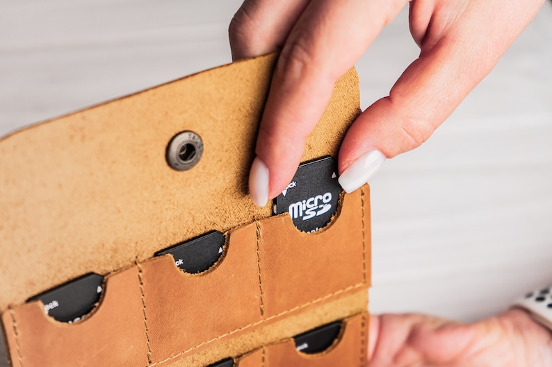 K&ouml;nnte beinhalten: Ein braunes Lederetui mit mehreren Steckpl&auml;tzen zum Aufbewahren von Speicherkarten. Eine Hand h&auml;lt eine Micro-SD-Karte mit der Aufschrift "micro SD" und steckt sie in einen der Steckpl&auml;tze.