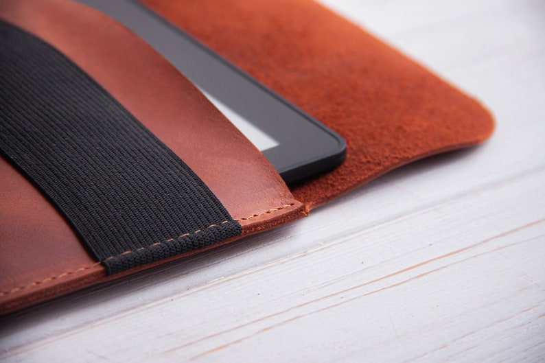 Puede incluir: Una funda de cuero marr&oacute;n para lector electr&oacute;nico con una correa el&aacute;stica negra. La funda est&aacute; cerrada y el lector electr&oacute;nico est&aacute; dentro.