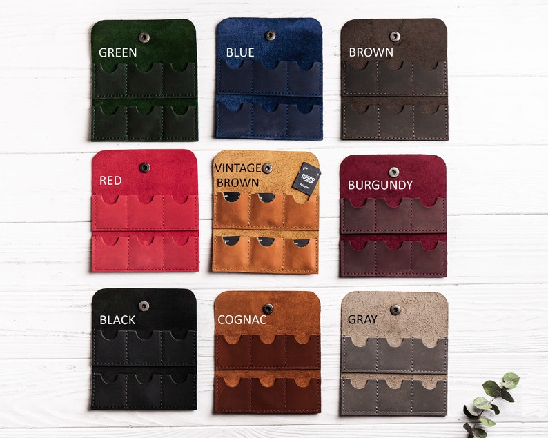 K&ouml;nnte beinhalten: Neun Lederh&uuml;llen f&uuml;r Speicherkarten in verschiedenen Farben, darunter Gr&uuml;n, Blau, Braun, Rot, Vintage Braun, Burgunderrot, Schwarz, Cognac und Grau. Jede H&uuml;lle hat acht Steckpl&auml;tze f&uuml;r Speicherkarten.