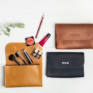 May include: An overhead shot of three leather cosmetic cases in brown and navy blue, with the names "CAMPER" and "DOLIE" embossed on two of them. A tan leather case is open, revealing makeup brushes, lipstick, and eyeshadow. A sprig of greenery is in the upper left.