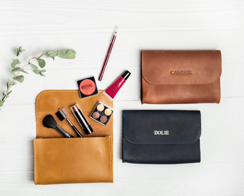 May include: An overhead shot of three leather cosmetic cases in brown and navy blue, with the names "CAMPER" and "DOLIE" embossed on two of them. A tan leather case is open, revealing makeup brushes, lipstick, and eyeshadow. A sprig of greenery is in the upper left.