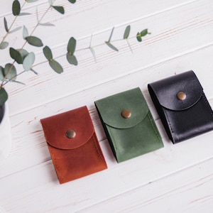 Pu&ograve; includere: Tre custodie in pelle marrone, verde e nera, ciascuna con chiusura a patta e bottone in ottone. Le custodie sono disposte su una superficie di legno bianco, con un piccolo vaso di verde e uno smartphone sullo sfondo.