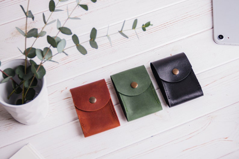 Pu&ograve; includere: Tre custodie in pelle marrone, verde e nera, ciascuna con chiusura a patta e bottone in ottone. Le custodie sono disposte su una superficie di legno bianco, con un piccolo vaso di verde e uno smartphone sullo sfondo.