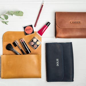 May include: An overhead shot of makeup and leather cosmetic bags. A mustard yellow leather bag is open, revealing makeup brushes, lipstick, and eyeshadow. A brown leather bag with "CAMBER" embossed on it and a navy blue bag with "DOLIE" are also present.